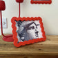 Small framed picture of a face on a wooden surface with a red lamp and 'London England' sign in the background.